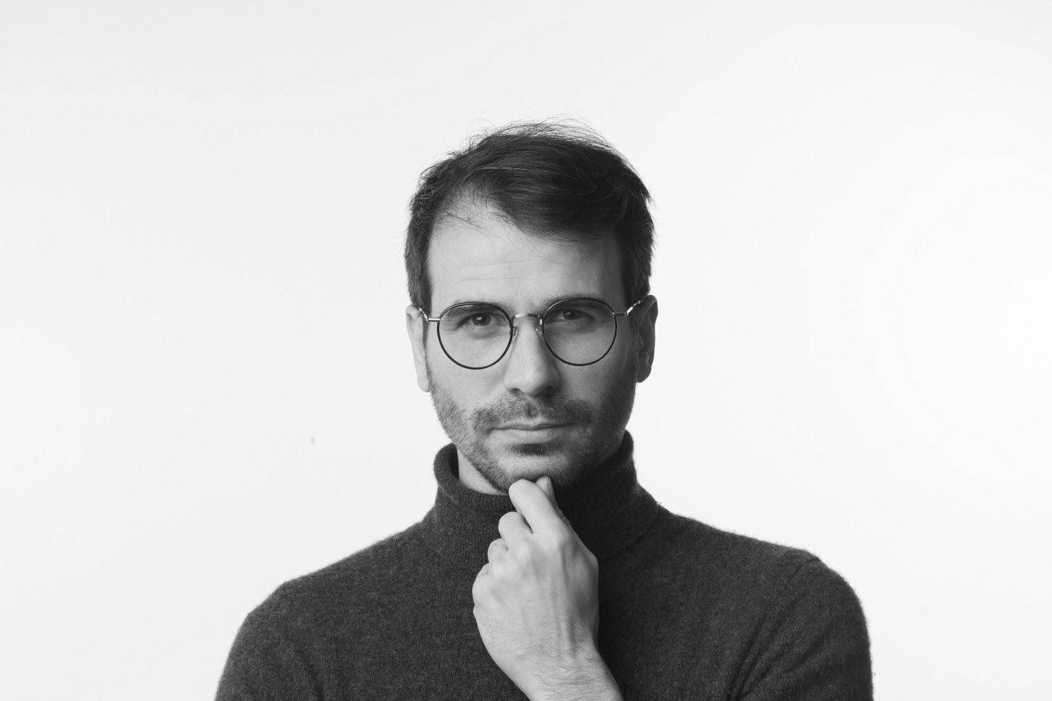Portrait en noir et blanc d’un homme en col roulé, lunettes rondes, main au menton